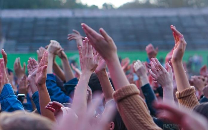 Audience with hands raised at a music festival and lights streaming down from above the stage.